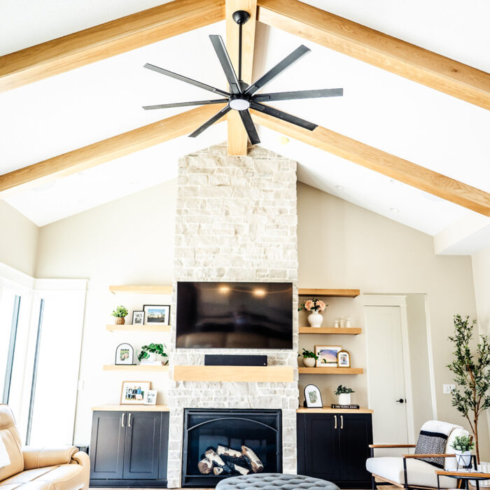 Bright living room with vaulted ceiling, exposed wooden beams, a large black ceiling fan, a stone fireplace, wall-mounted TV, built-in shelves with décor, and modern furniture. Large windows let in natural light in remodeled home in Huxley Iowa