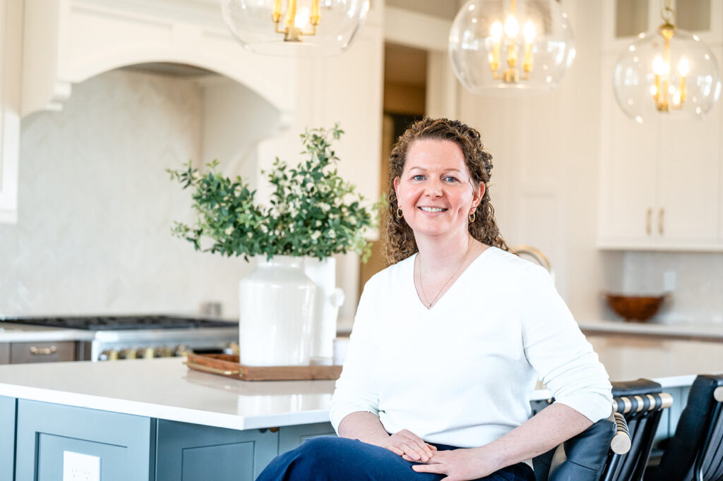 Karen Roa of Reese Builders wearing a white top and navy pants, sits smiling on a stool in a bright, modern remodeled kitchen in Huxley Iowa with white cabinets, gold light fixtures, and a vase of greenery on the island.