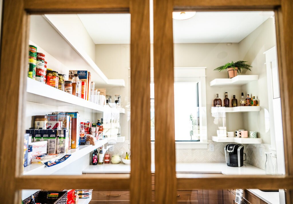 A well-organized pantry with white shelves holding canned goods, snacks, bottles, a coffee maker, and a potted plant, viewed through glass-paned wooden doors.