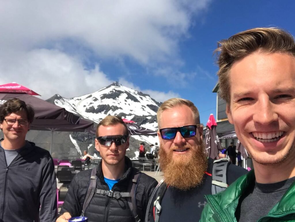 Four men pose for a selfie outdoors at a mountain resort, with snow-capped peaks and blue sky in the background. They wear jackets and sunglasses, and outdoor seating with umbrellas is visible behind them.