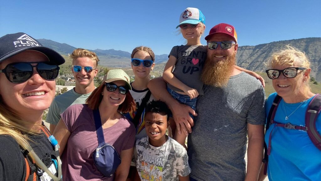 A group of eight people, including adults and children, smile at the camera outdoors with mountains in the background on a sunny day. They wear casual clothes, sunglasses, and hats.