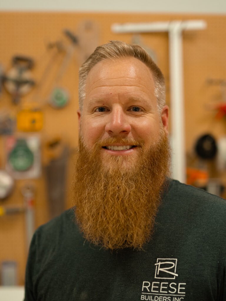 A man with a long red beard and short blond hair smiles, standing in front of a pegboard wall with various tools hanging. He wears a dark green "Reese Builders Inc." T-shirt.