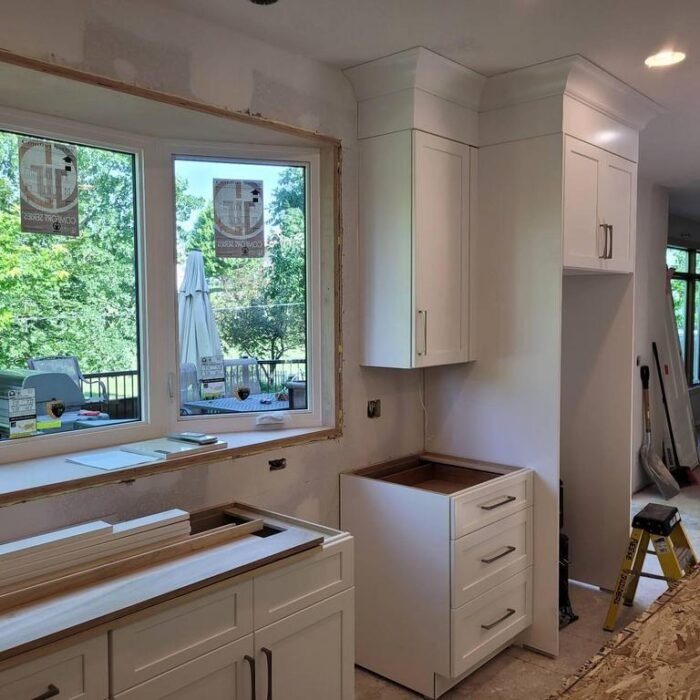 A kitchen in Des Moines under renovation for a remodel features white cabinets, an unfinished countertop, a ladder, and unpainted window trim. Large windows reveal Iowa greenery outside, with building materials scattered around the room.