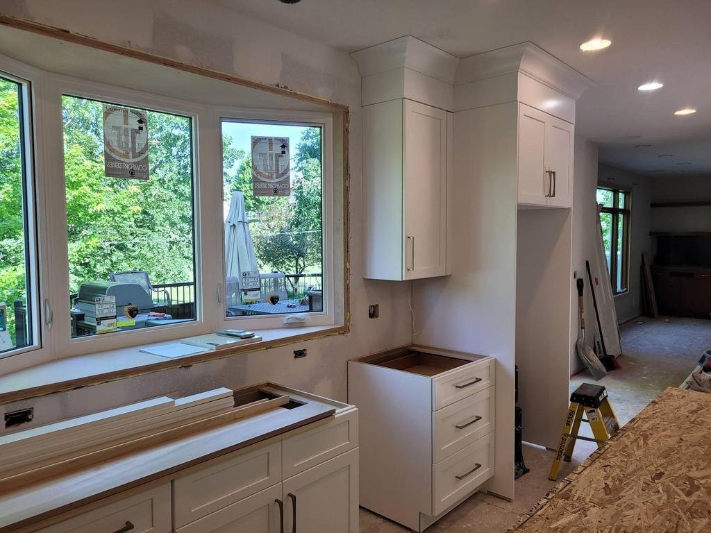 A kitchen in Des Moines under renovation for a remodel features white cabinets, an unfinished countertop, a ladder, and unpainted window trim. Large windows reveal Iowa greenery outside, with building materials scattered around the room.