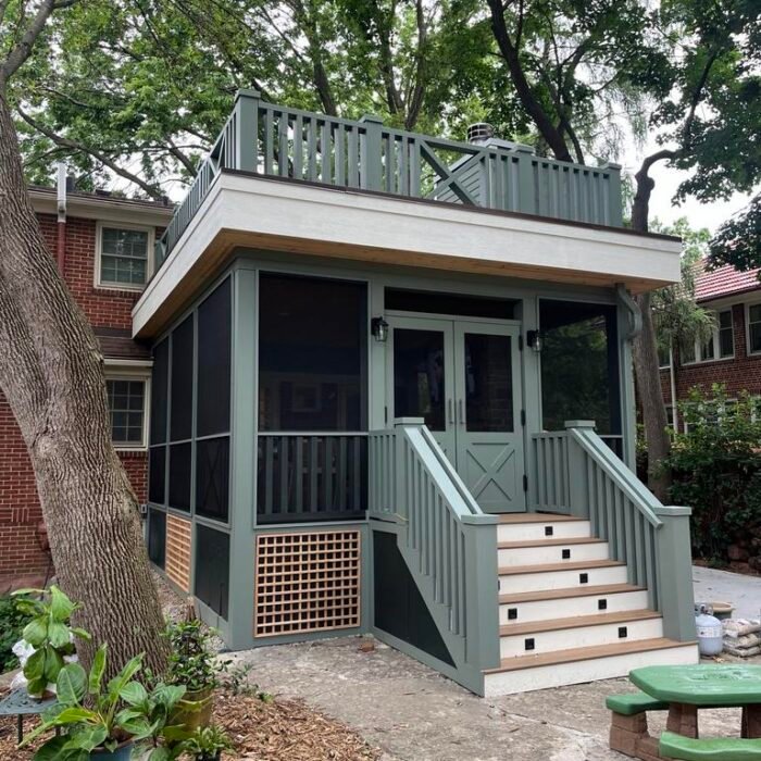 A two-story screened porch with green and white trim offers a charming backyard retreat in Des Moines, featuring stairs to a covered entrance, a rooftop deck, and lush trees—perfect inspiration for your next renovation.