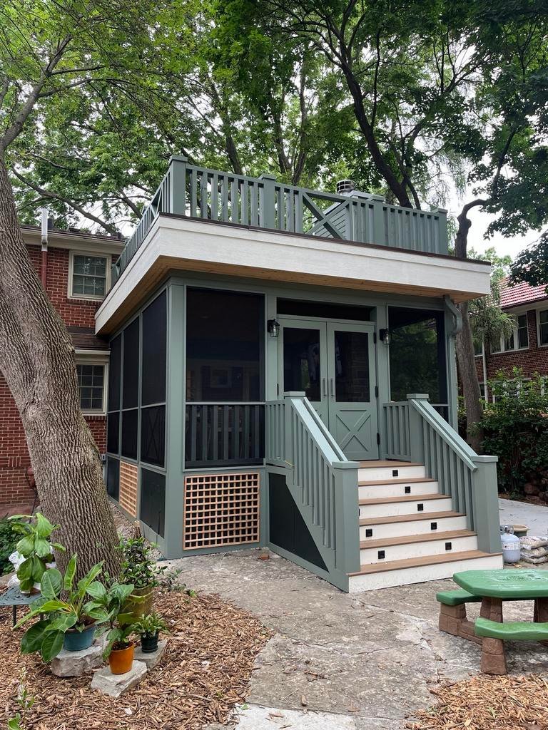 A two-story screened porch with green and white trim offers a charming backyard retreat in Des Moines, featuring stairs to a covered entrance, a rooftop deck, and lush trees—perfect inspiration for your next renovation.