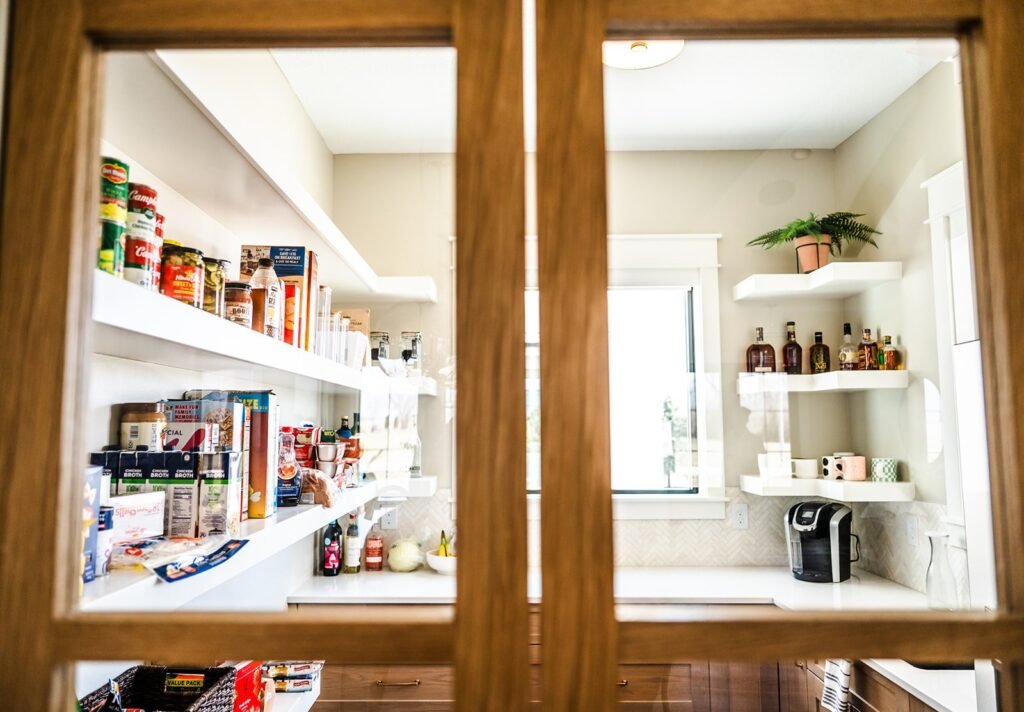View through glass doors into a bright, organized pantry with white shelves—an example of smart design for any Iowa home addition—stocked with canned goods, snacks, drinks, kitchen supplies, and a coffee maker under the window. A plant sits on an upper shelf.