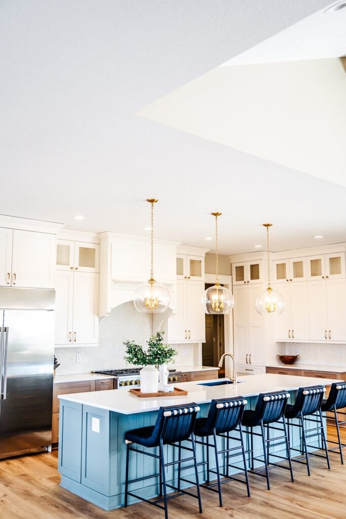 A modern Des Moines kitchen featuring white cabinets, a large stainless steel fridge, a light blue island with a white countertop, five black barstools, and three glass pendant lights completing this stunning remodel.