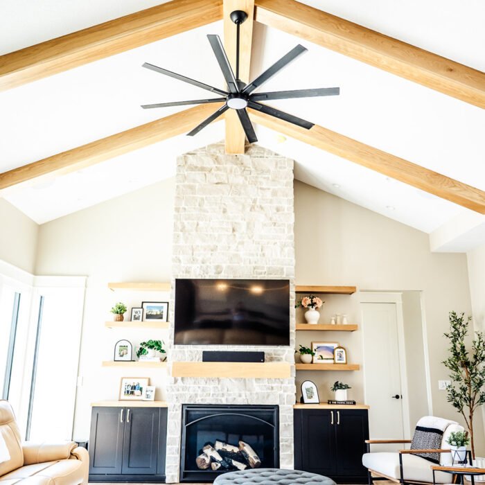 Bright living room in this Des Moines, Iowa home with vaulted ceiling, exposed wooden beams, a large black ceiling fan, stone fireplace with TV above, built-in cabinets and shelves, modern furniture, and large windows flooding the addition with natural light.