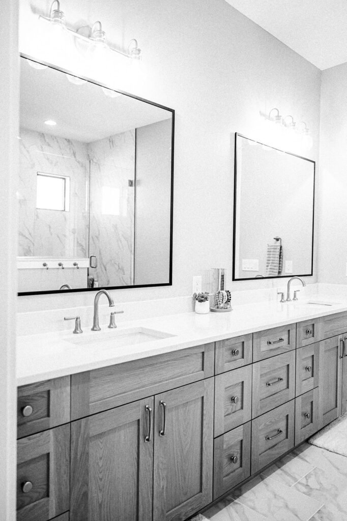 A modern bathroom in Des Moines featuring a double vanity, light wood cabinets, two rectangular mirrors, and dual sinks. The space showcases marble floor and wall tiles, with bright lighting above each mirror—perfect for your next remodeling project.