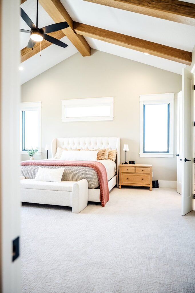 Bright, modern bedroom in a renovated Des Moines, Iowa home with vaulted ceiling, exposed wooden beams, ceiling fan, tufted bed, cozy bedding, a bench at the foot, wooden nightstands, and large windows letting in natural light.