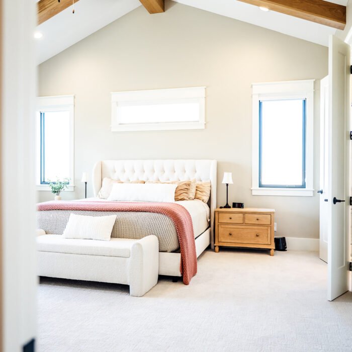 Bright, modern bedroom in a renovated Des Moines, Iowa home with vaulted ceiling, exposed wooden beams, ceiling fan, tufted bed, cozy bedding, a bench at the foot, wooden nightstands, and large windows letting in natural light.