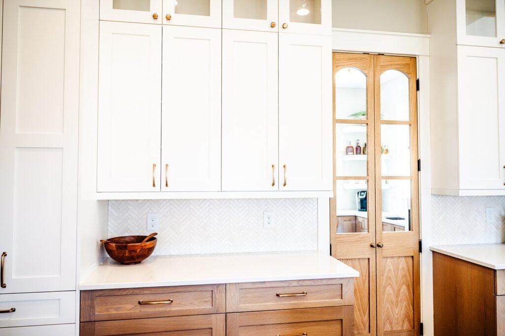 Bright, modern kitchen in a Des Moines home with white upper cabinets, light wood lower cabinets, brass handles, a white backsplash, a wooden bowl on the counter, and glass-paneled wooden doors showcasing fine, custom carpentry leading to a pantry.