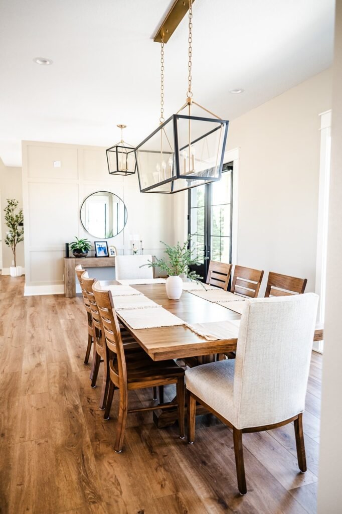 Bright, modern dining room connects seamlessly to the kitchen, featuring a long wooden table, mixed chairs, large pendant lights, potted centerpiece, and hardwood floors. Wall art and plants enhance the airy feel—perfect for a small remodel in Iowa.