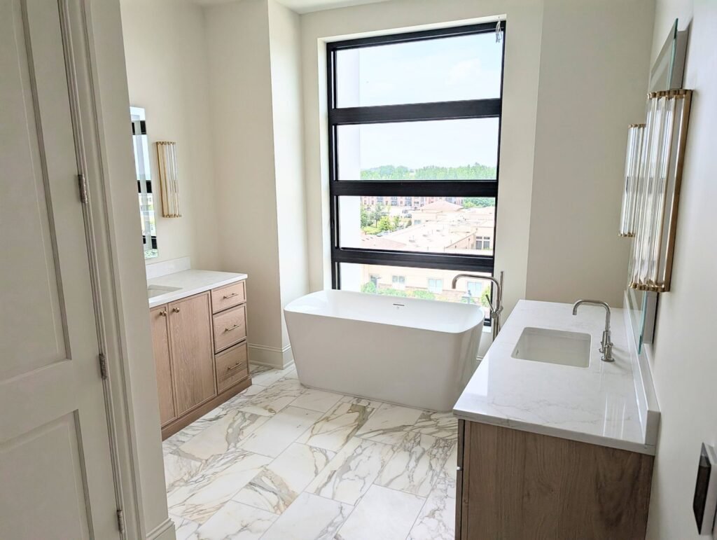 Modern bathroom remodel in Des Moines featuring a large window with city views, freestanding white bathtub, light wood cabinets, marble countertops, and marble tile floor. The space is brightened by natural light throughout.