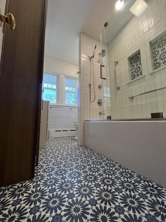 Bathroom Remodel in Des Moines featuring a patterned black and white tile floor, white tiled shower with glass door, built-in shower niches, chrome fixtures, and three large windows for natural light.