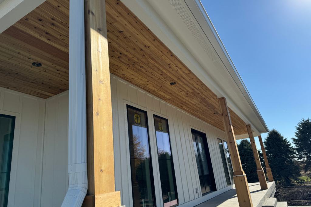 A low-angle view of a house porch with wooden posts and a wood-paneled ceiling. The house has light-colored siding, large windows, and a sloped roof. Trees and a clear blue sky are visible in the background.