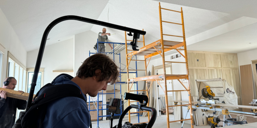 A camera operator films inside a house under construction. Two workers stand on scaffolding, one painting near the ceiling, while another carries wood. Tools, ladders, and construction materials fill the room.