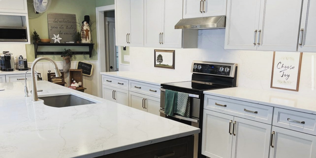 Bright modern kitchen with white cabinets, marble countertops, stainless steel appliances, and a large island with a sink. Decorative signs and a small snowman figure are displayed on shelves and countertops.