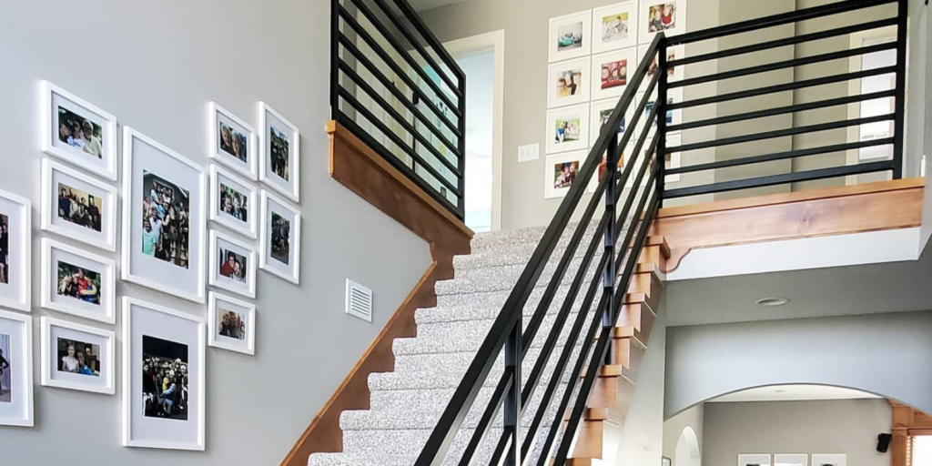 A carpeted staircase with black metal railings and wooden trim ascends beside a wall decorated with a gallery of framed family photos in a modern, light-filled home interior.