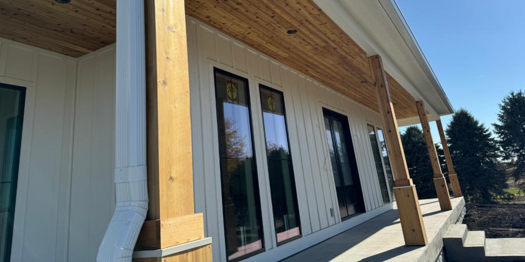A close-up angled view of a house’s covered porch, featuring wooden columns, white siding, vertical windows with transoms, and a wood-paneled ceiling, with trees visible in the background.